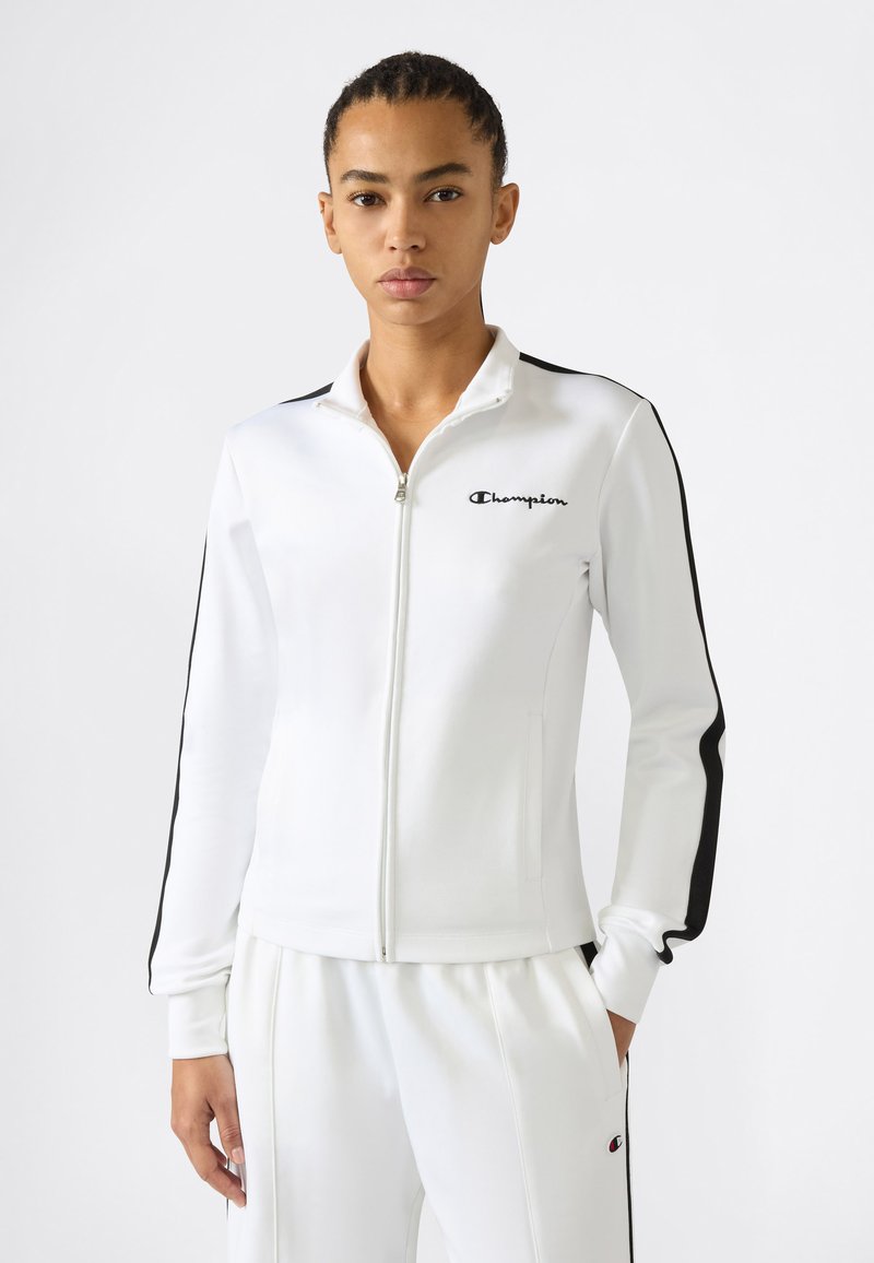 Femme portant une veste de sport blanche zippée et un pantalon assorti avec des rayures noires et le logo Champion sur la poitrine et la cuisse.