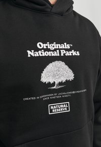Schwarzer Kapuzenpullover mit weißem Text und einer Grafik eines Baumes. Merkmale umfassen eine Kängurutasche und weichen Stoff. Der Text lautet "Originals National Parks."