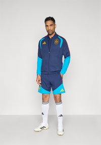 Conjunto de entrenamiento que incluye una chaqueta azul marino con detalles en aguamarina y rojo, pantalones cortos a juego en azul marino con detalles en aguamarina, calcetines blancos con rayas negras y botas de fútbol.