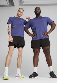 To männliche Figuren står side by side, iført lilla atletiske t-shirts og sorte shorts. Fodtøjet har kontrasterende farver: hvidt med gule accenter og sort.