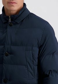Homme portant une veste d'hiver matelassée bleu foncé avec des boutons noirs et une fermeture éclair, montré de la nuque à mi-torse sur un fond clair.