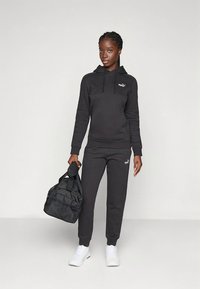 Czarna bluza z kapturem i spodnie dresowe wykonane z miękkiego materiału, z małym białym logo. Zestaw uzupełnia czarna torba typu duffel oraz białe sneakersy.