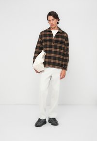 Brown and black plaid shirt with button-up front, two chest pockets, white t-shirt, light-colored trousers, and dark sneakers. Holding a cream bag.