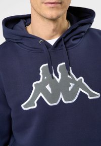 Sudadera de color azul marino en mezcla de algodón, con un gran logo bordado en gris que muestra a dos figuras sentadas. Incluye capucha con cordones.