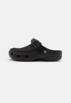 Zueco negro de tipo slip-on con correa ajustable, suela perforada y logotipo circular en el lateral, diseñado para uso casual y comodidad.