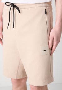 Beige sporta šorti ar auklu viduklī, sānu kabatām un melnu logo uzlīmi. Izgatavoti no gludas auduma, ar brīvu piegriezumu.