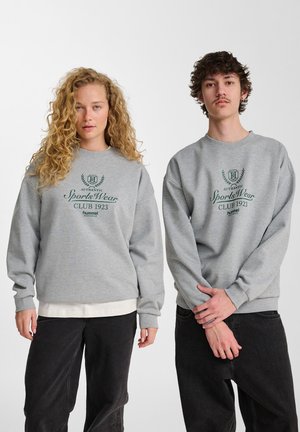 Les sweatshirts à col rond gris présentent un texte et des logos imprimés en vert, avec des poignets et un ourlet côtelés. Porté avec un pantalon en denim foncé.