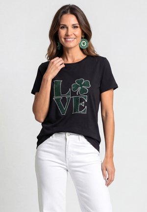 Uśmiechnięta kobieta nosząca czarny t-shirt z "LOVE" oraz zielonym motywem koniczyny, w połączeniu z białymi spodniami i dużymi, okrągłymi zielonymi kolczykami.