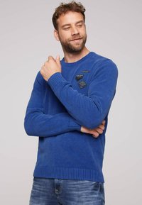 Maglione blu a maglia con polsini e orlo a costine, caratterizzato da tre toppe di tessuto sul petto. Indossato con jeans blu, sfondo semplice.