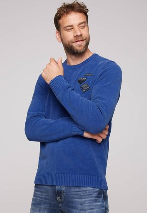 Blauwe gebreide trui met geribde manchetten en zoom, versierd met drie stofpatches op de borst. Draagt met blauwe spijkerbroek, op een effen achtergrond.