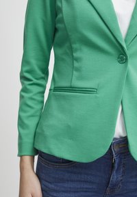 Zelený blazer s jedním knoflíkem, přiléhavý střih, hladká textura a boční kapsa; nošený přes bílé tričko a modré džíny.