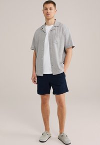 Szürke, rövid ujjú ing, fehér, kerek nyakú póló alatta, tengerészkék rövidnadrág és szürke slip-on cipő. Tartalmaz egy mellzsebet és gombokat.