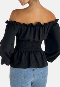 Sort off-the-shoulder bluse med flæsset halsudskæring og smocket talje. Lange, puffede ærmer og letvægtsstof. Båret med blå jeans.