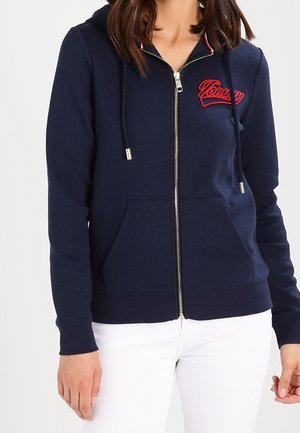 Femme portant un sweat à capuche zippé bleu marine avec un logo "Tommy" rouge sur la poitrine et un pantalon blanc, debout devant un fond uni.