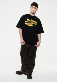 Black t-shirt featuring "The Beatles Yellow Submarine" graphic in yellow and red. The model wears loose, dark brown pants and black shoes.