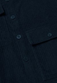 Dettaglio ravvicinato di tessuto in corduroy blu scuro con righe verticali, dotato di quattro bottoni neri e una tasca a patta.