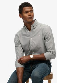 Camisa gris claro, de botones y de mangas largas, con un pequeño logo en el pecho. Lleva puesta con unos pantalones vaqueros azul oscuro y un reloj dorado.