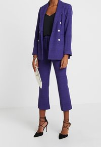Completo a scacchi viola con blazer doppiopetto e pantaloni cropped, abbinato a una maglia nera, décolleté neri e pochette bianca.
