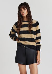 Giovane donna con lunghi capelli castani che indossa un maglione a righe beige e nere e pantaloni corti in pelle nera, in piedi contro uno sfondo neutro.