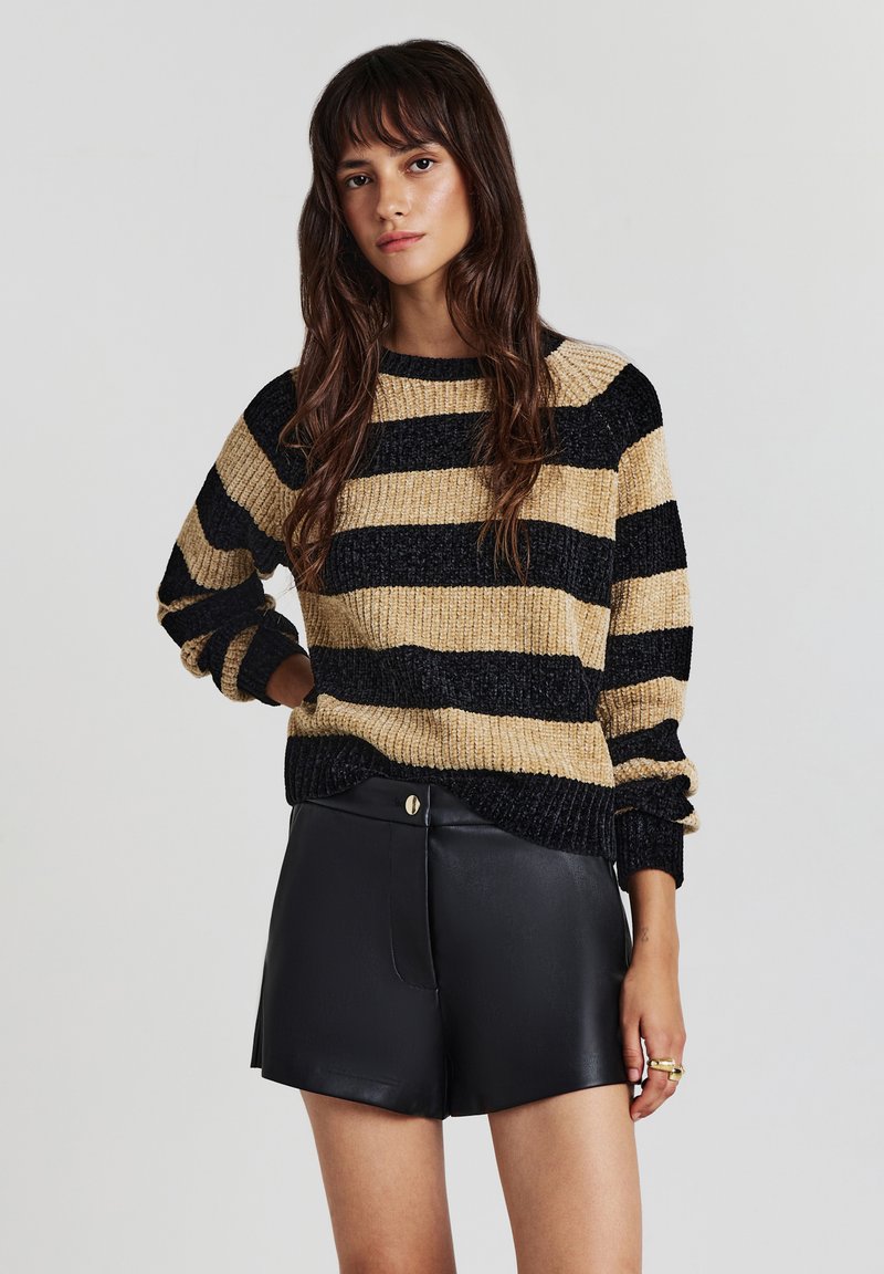 Giovane donna con lunghi capelli castani che indossa un maglione a righe beige e nere e pantaloni corti in pelle nera, in piedi contro uno sfondo neutro.