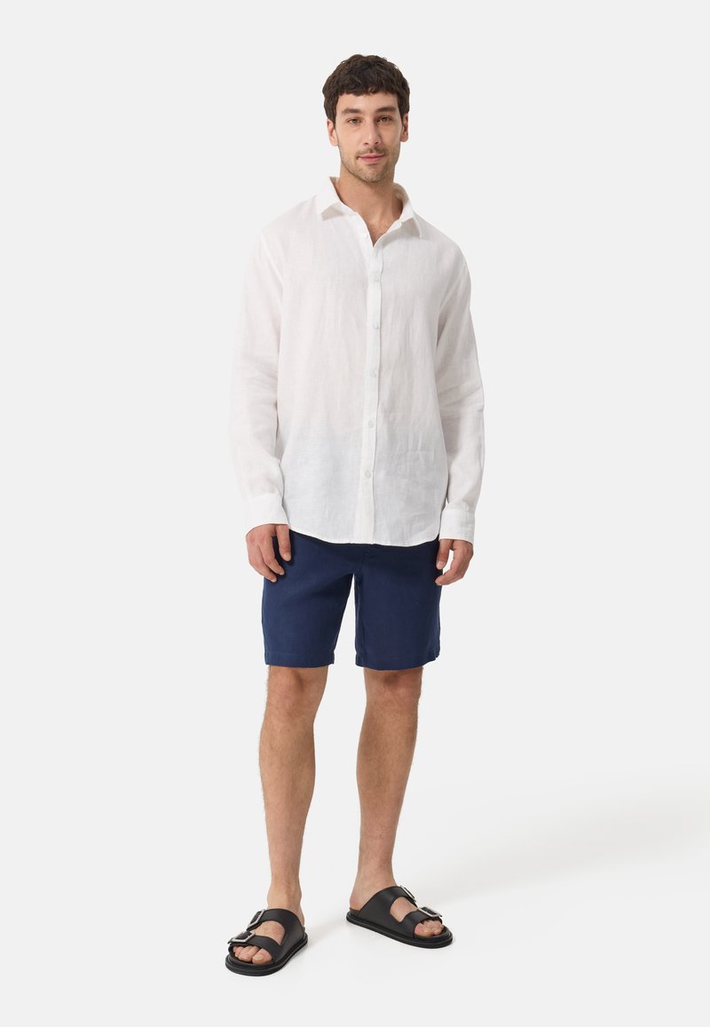 Chemise en lin blanche avec un devant boutonné, shorts bleu marine, et sandales noires avec un détail de boucle en argent. Design simple et coupe décontractée.