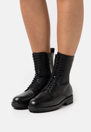 Un par de botas de cuero negro de cordones hasta el tobillo con cremalleras laterales, llevadas sobre piernas desnudas, contra un fondo blanco liso.