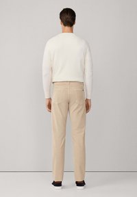 Mann, der einen cremfarbenen Pullover, beige Hose und schwarze Sneaker trägt, steht vor einer leeren hellgrauen Wand.