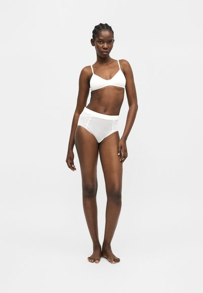 Γυναίκα στέκεται ξυπόλητη, φοράει λευκό bralette και εσώρουχα από δίχτυ με ψηλή μέση σε απλό ανοιχτό γκρι φόντο.