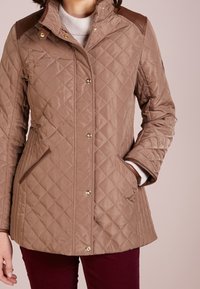 Prošívaná hnědá bunda se zlatými detaily, hnědými ramenními akcenty, bočními kapsami a vysokým límcem. Texturovaný materiál s hladkým povrchem.