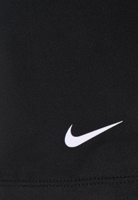 Logo Nike swoosh blanc sur tissu noir texturé avec des coutures visibles près du bord inférieur.
