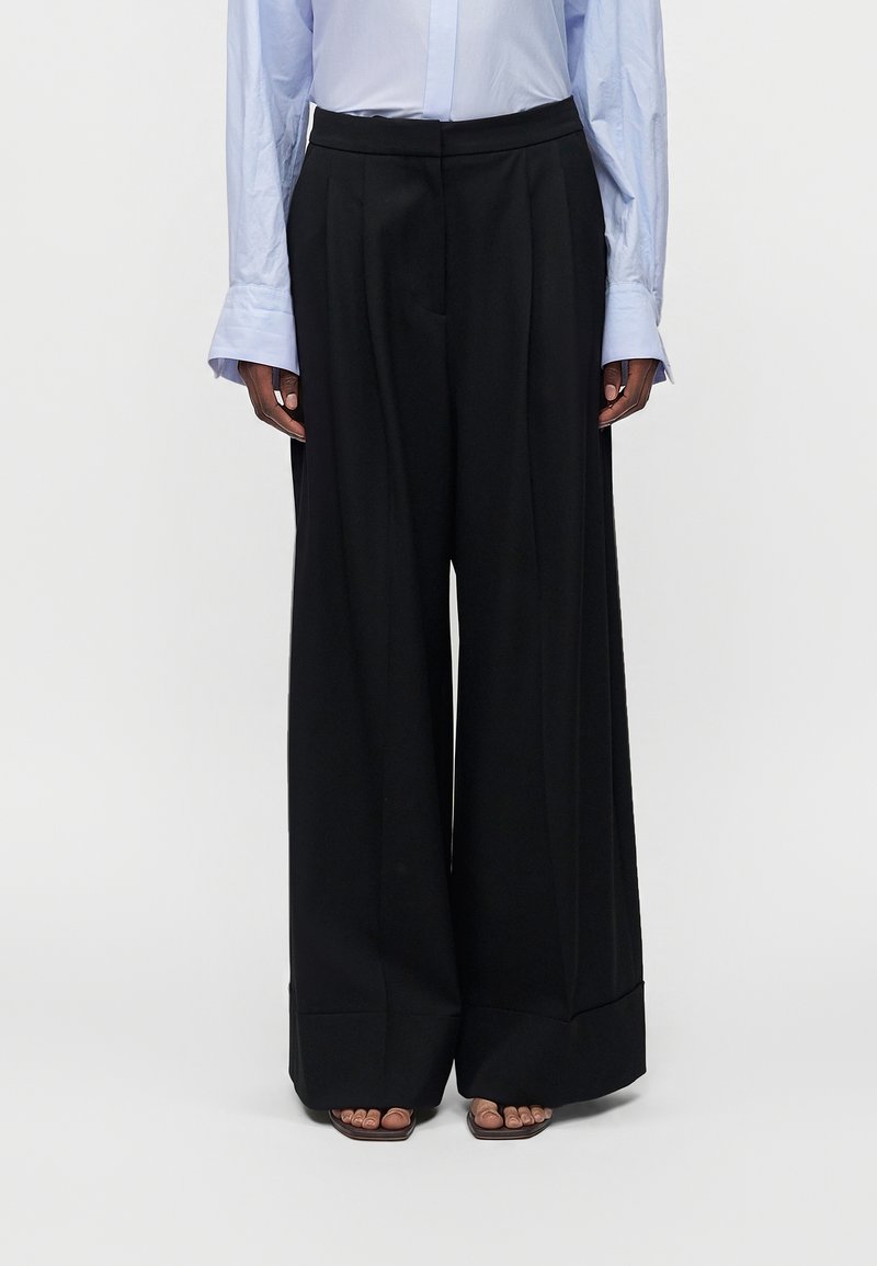 Personne portant un pantalon large noir, une chemise bleu clair à manches longues et des sandales marron à bouts ouverts, debout devant un fond blanc.