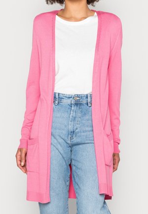 Person iført en lang lyserød cardigan over en hvid t-shirt og lyseblå højtaljede jeans, stående foran en ensfarvet baggrund.