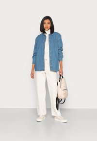 Denimskjorte i blått, lange ermer, knapping foran, båret over en hvit turtleneck. Kombinert med hvite vide bukser og beige joggesko, holder en ryggsekk.