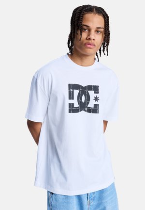 Jongere met gevlochten haar, gekleed in een wit T-shirt met een zwart DC-logo en blauwe spijkerbroek, staand tegen een effen achtergrond.