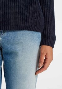 Pull en maille côtelée bleu marine à manches longues, associé à un jean denim bleu clair. Gros plan sur une main posée sur le jean.