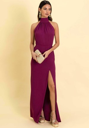 Robe longue et profonde couleur violet avec un décolleté halter et une fente haute sur la cuisse. Le mannequin tient une pochette beige et porte des talons compensés argentés.