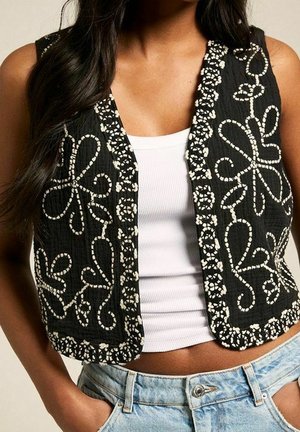 Sleeveless black vest with white floral embroidery worn over a white ribbed tank top and light blue denim jeans.