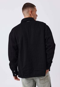 Schwarze strukturierte Jacke mit Fischgrätmuster, ausgestattet mit einem Kragen und knöpfbaren Manschetten. Das Modell wird von hinten gezeigt und trägt helle Jeans.