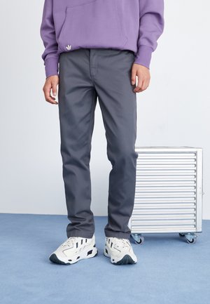 Une personne portant un pantalon gris, des baskets blanches avec des rayures noires, et un sweat à capuche violet se tenant à côté d'une valise à roulettes argentée.
