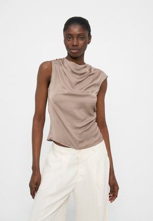 Naine, kes kannab varrukateta savipunase tooniga lipsuga ülevalpool olevat paelaga pluusi ja kõrge vöökohaga valgeid pükse, seisab ühevärvilise heleda tausta ees.