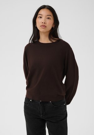Femme aux longs cheveux foncés portant de petites boucles d'oreilles créoles, un pull marron foncé et un jean noir, debout devant un fond clair.