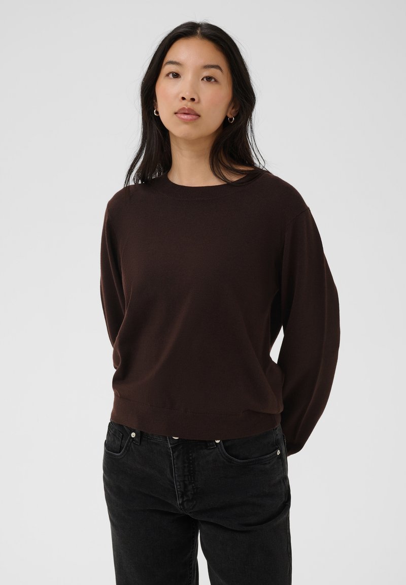 Femme aux longs cheveux foncés portant de petites boucles d'oreilles créoles, un pull marron foncé et un jean noir, debout devant un fond clair.