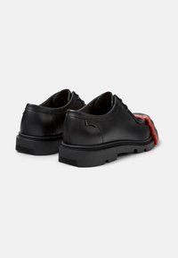 Zapatos de cuero negro con punta redondeada, que presentan un diseño distintivo en rojo y negro en la suela exterior, suela de goma texturizada y un sutil logotipo.