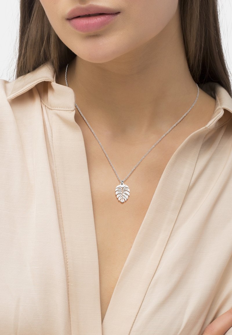 Femme portant un collier chaîne argenté avec un pendentif en forme de feuille détaillée sur un chemisier beige à col ouvert.