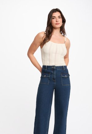 Jeune femme aux longs cheveux foncés portant un haut sans manches en maille crème et un jean bleu taille haute à jambes larges avec des poches à rabat à l'avant.