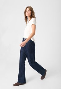 Jean en denim évasés avec finition bleu foncé, dotés de cinq poches et de détails cousus, associés à un T-shirt uni de couleur claire et des chaussures marron.
