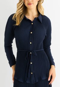 Navy blue pleated blouse with long sleeves, button front, and tie waist. Features gold buttons and textured fabric for added detail.