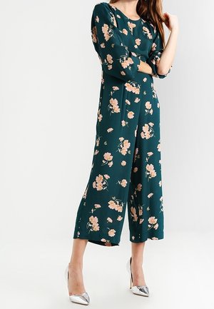 Femme portant une combinaison pantalon large à motifs floraux bleu sarcelle avec manches trois-quarts et talons hauts argentés pointus, debout sur fond blanc.