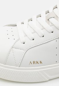 Witte leren sneaker met een gouden "ARKK" logo, gestructureerde witte rubberen zool, vlakke witte veters en geperforeerde details.
