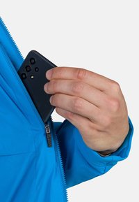 Sininen takki, jossa on vetoketjullinen tasku, jossa pidetään mustaa älypuhelinta. Käsi ulottuu ottamaan puhelimen, joka esittelee taskun muotoilua.